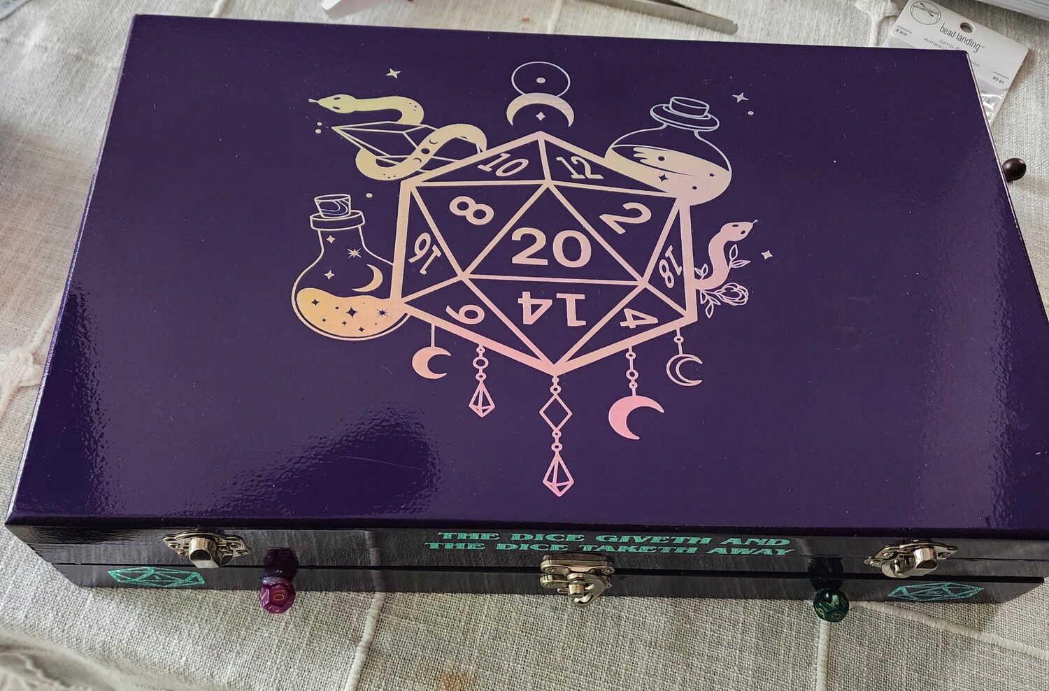 Violet dice box lid with a holographic d20 crest ringed by snakes, crescent moons and potion bottles